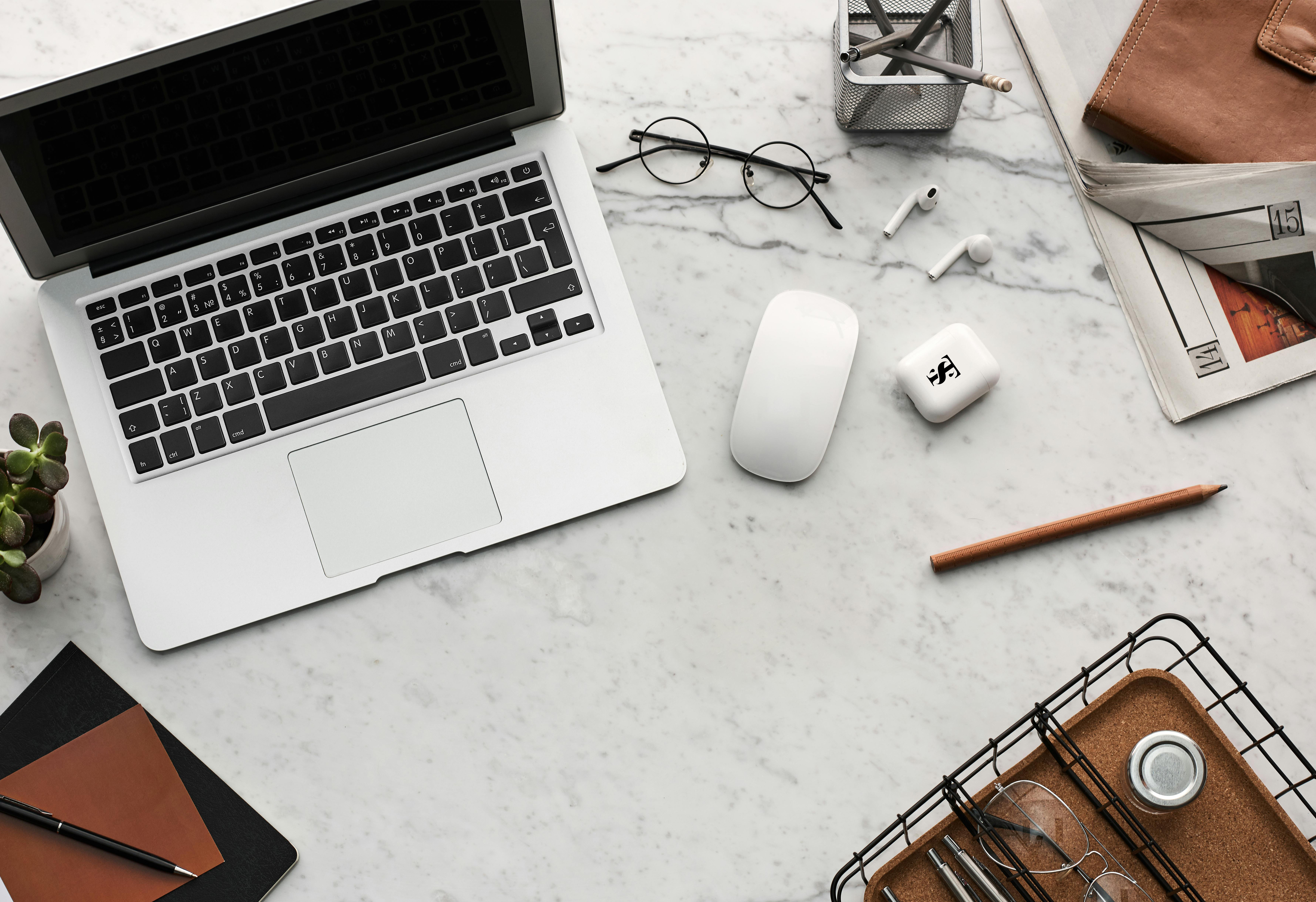 Top-down view of a modern workspace featuring a laptop, wireless earbuds, eyeglasses, notebook, pencil, and desk accessories on a white marble surface
